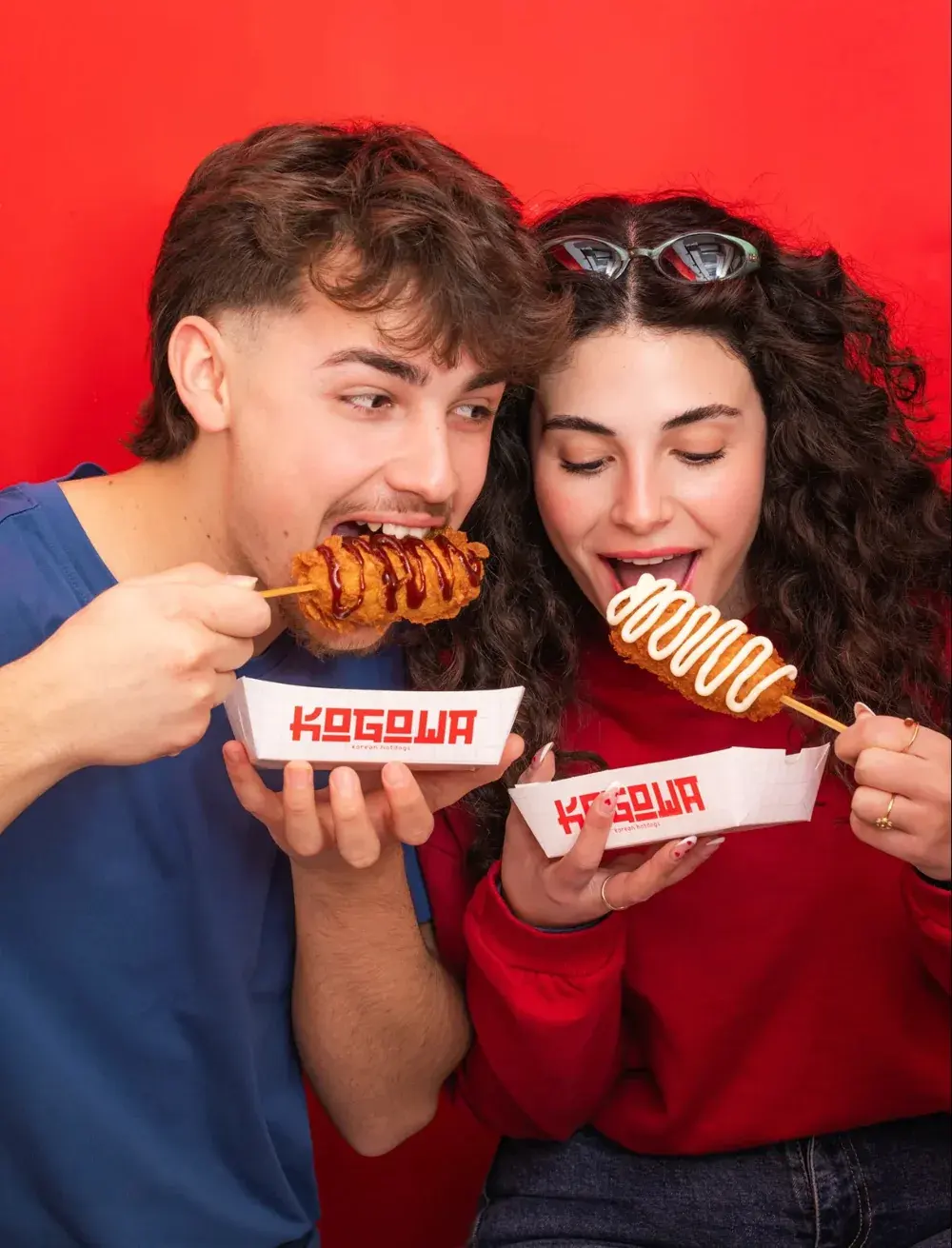 Two friends biting into Kogowa hotdogs on sticks, one with BBQ sauce and one with mayo, holding Kogowa trays against a red wall