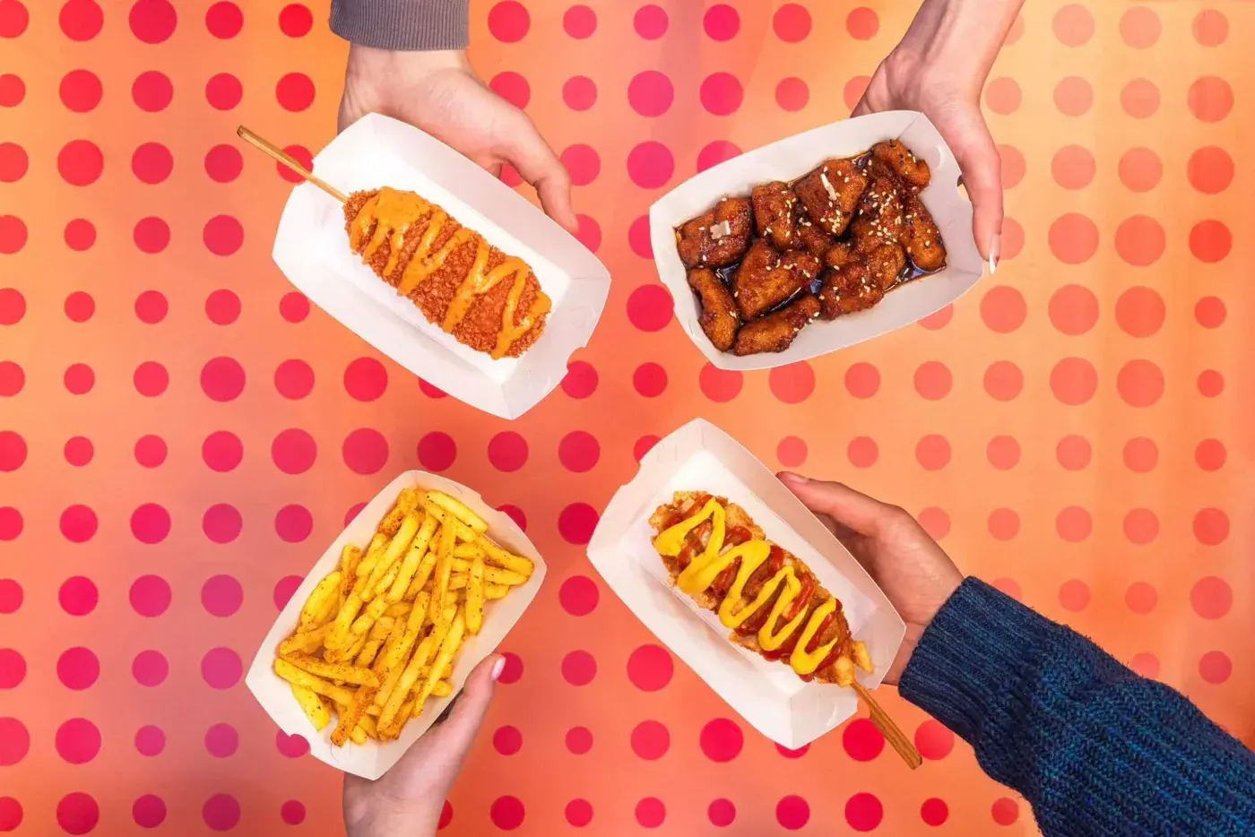 Four hands holding Kogowa trays: two hotdogs on sticks with sauce, sesame chicken and seasoned fries, over a pink dotted table
